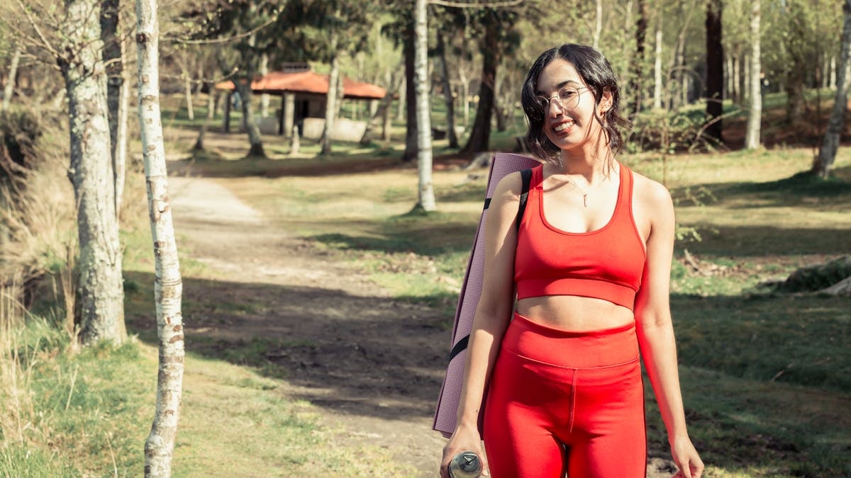 Smiling woman in red sport clothing holding a yoga mat outdoors.